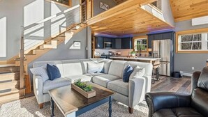 Un salón amplio con un sofá seccional gris, una mesa de centro de madera y un sillón de cuero negro. Una escalera con barandilla de madera conduce a un nivel superior.