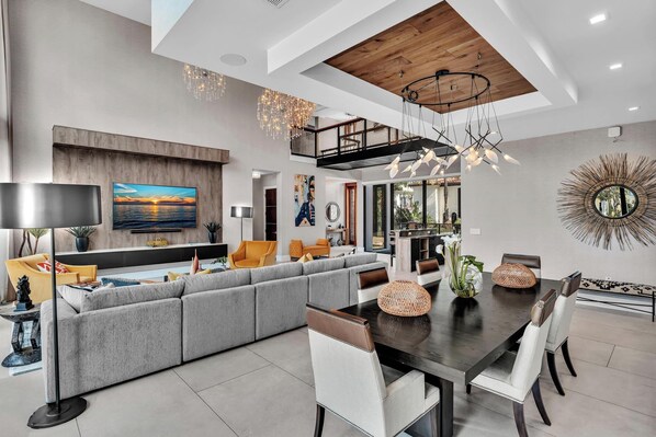 A modern living room with a sectional sofa, dining table, and chairs. There is a large painting on the wall, a chandelier, and a decorative mirror. - Coral Heaven | Las Olas Waterfront Estate | Dock (Fort Lauderdale)