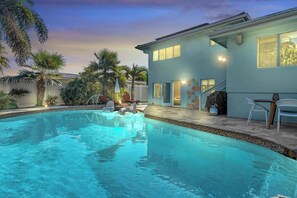 Uma piscina com espreguiçadeiras, um alpendre coberto e uma casa com fachada azul clara.