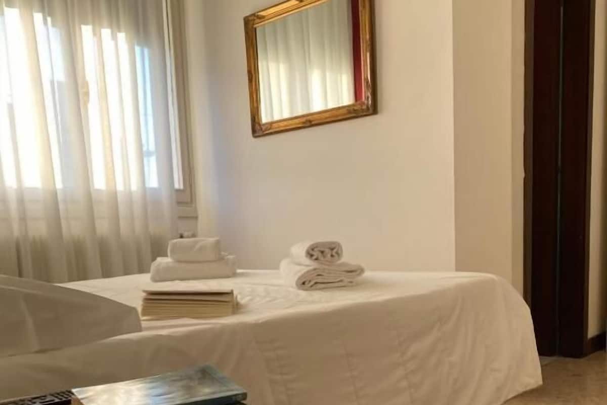 Ein Hotelzimmer mit Bett, Nachttisch mit Büchern, gefaltetem Handtuch und Spiegel an der Wand.
