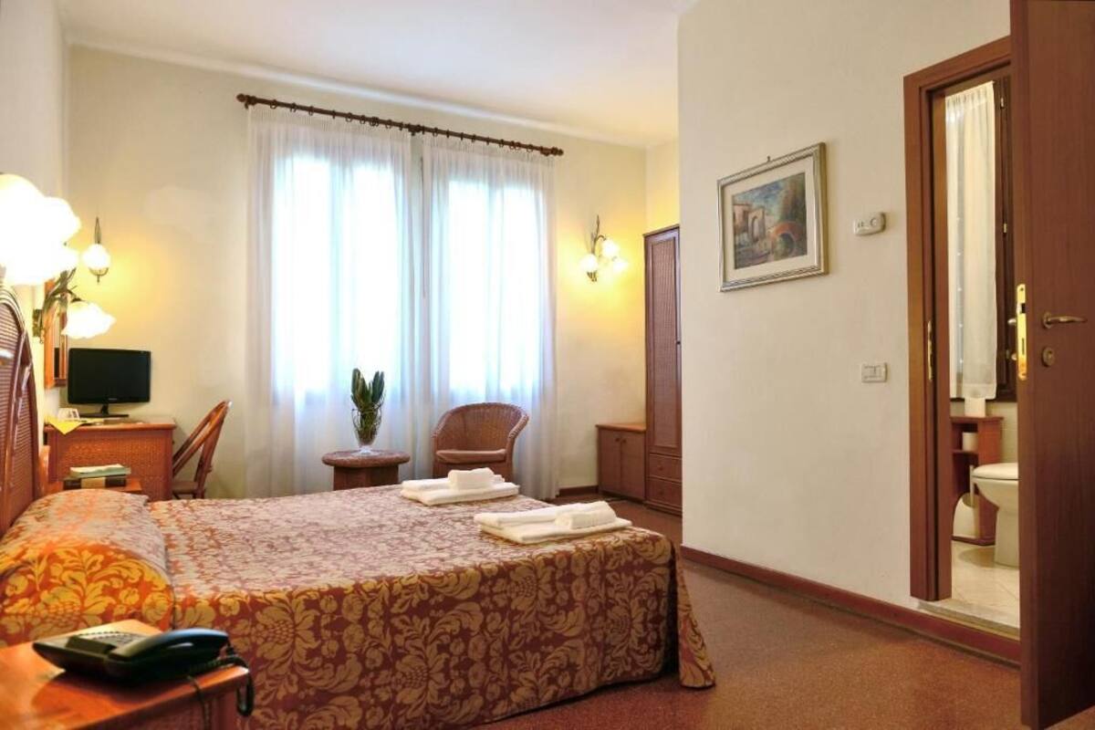 Ein Hotelzimmer mit einem Bett, einem Schreibtisch mit Telefon, einem Sessel, einem kleinen Tisch mit Pflanze und einem Bild an der Wand.