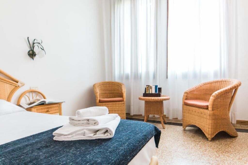 Een hotelkamer met een bed, twee rieten stoelen, een klein tafeltje met boeken en een raam met dunne gordijnen.
