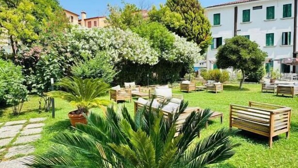 A garden with outdoor seating, a palm tree, and a building in the background. - Hotel Orio (Venice)