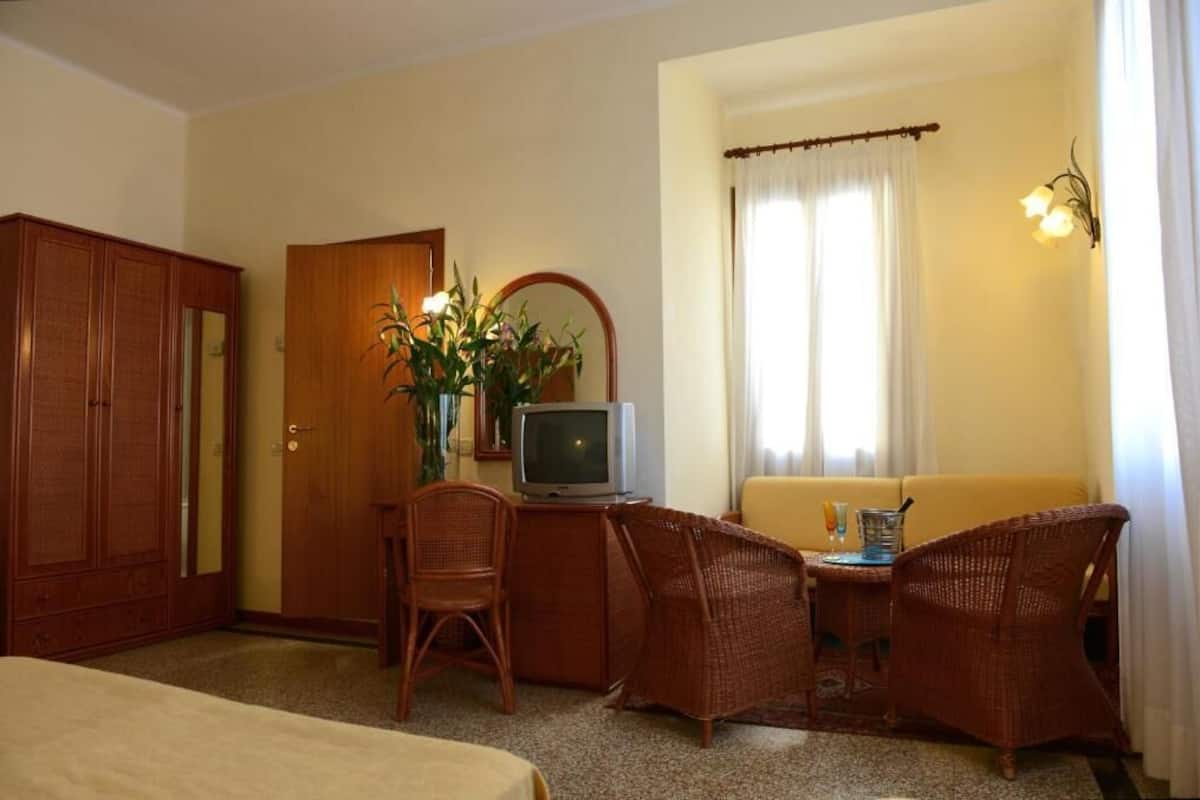 Ein Hotelzimmer mit einem Bett, einem Schreibtisch mit Fernseher, Korbstühlen, einer Couch und einem Fenster mit Vorhängen.