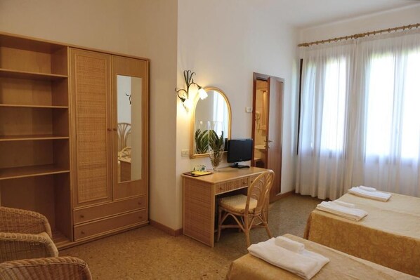 Habitación de hotel con armario de madera, escritorio con computadora, silla, cama con dos almohadas y ventana con cortinas.