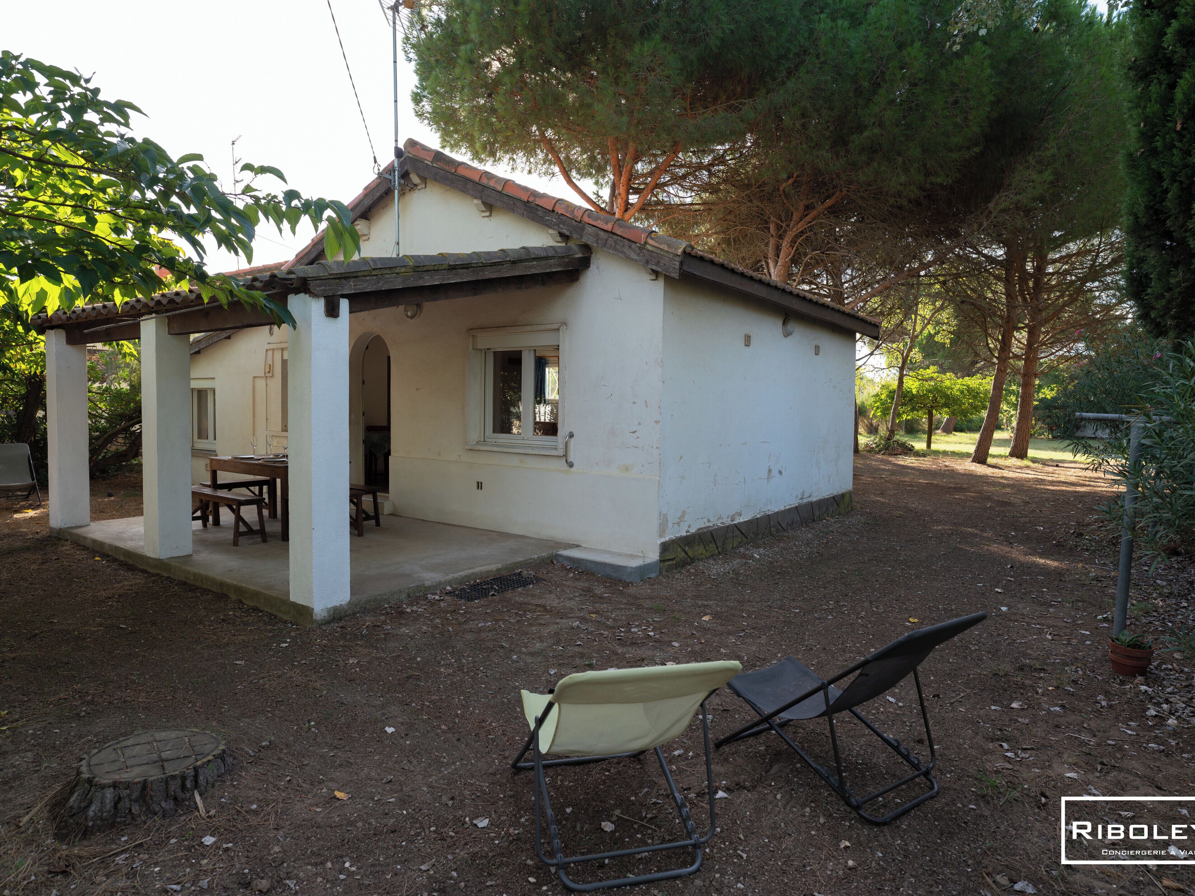 A small white house with a tiled roof, a patio with a table and chairs, and a garden with trees and a bench.