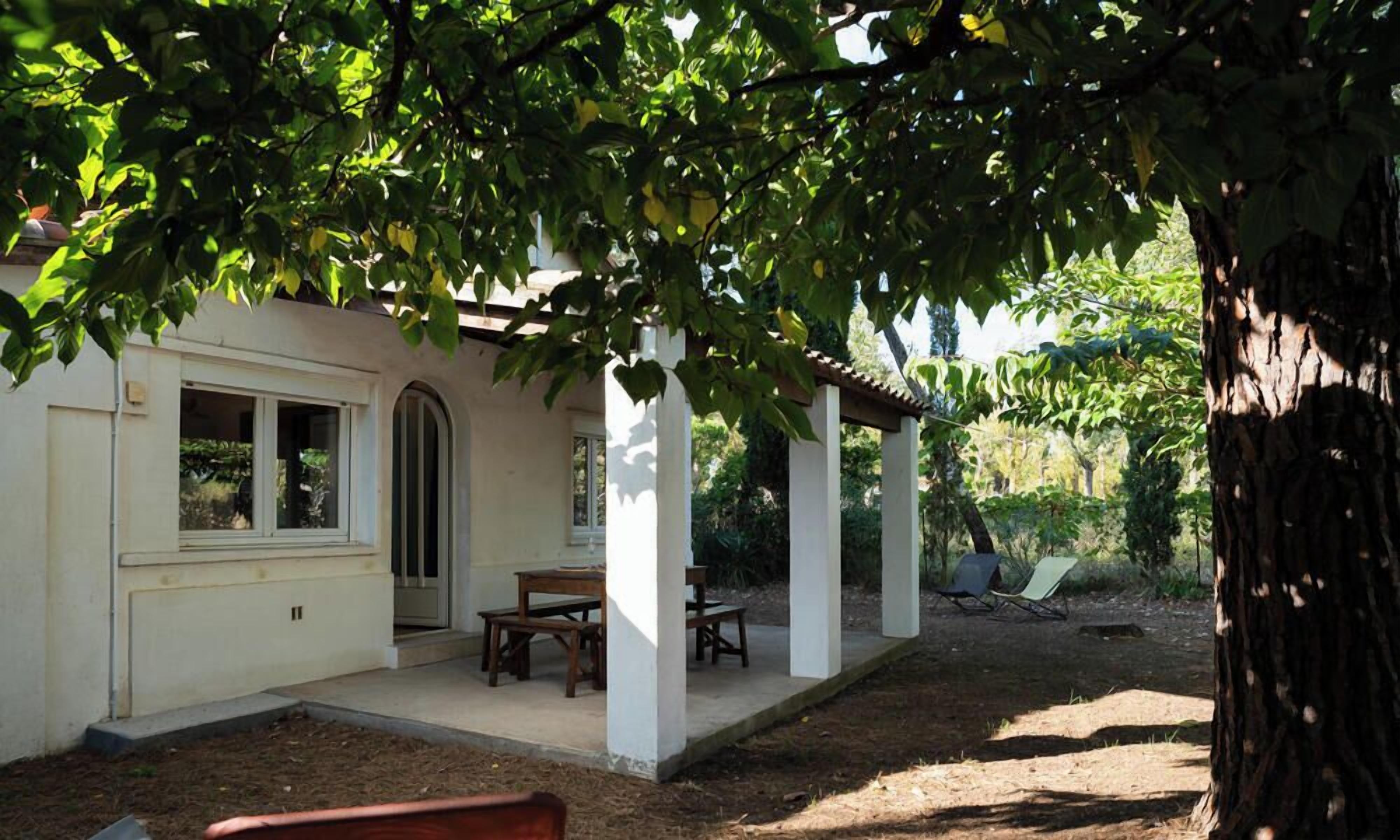 Ein überdachter Patio-Bereich mit Tisch und Stühlen, ein weißes Gebäude mit Fenster und ein Baum im Vordergrund.