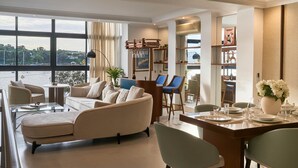 Un salon moderne avec un canapé, une table basse et des fauteuils. On y trouve également un coin repas avec une table dressée pour deux personnes. Une grande fenêtre offre une vue sur l’extérieur.