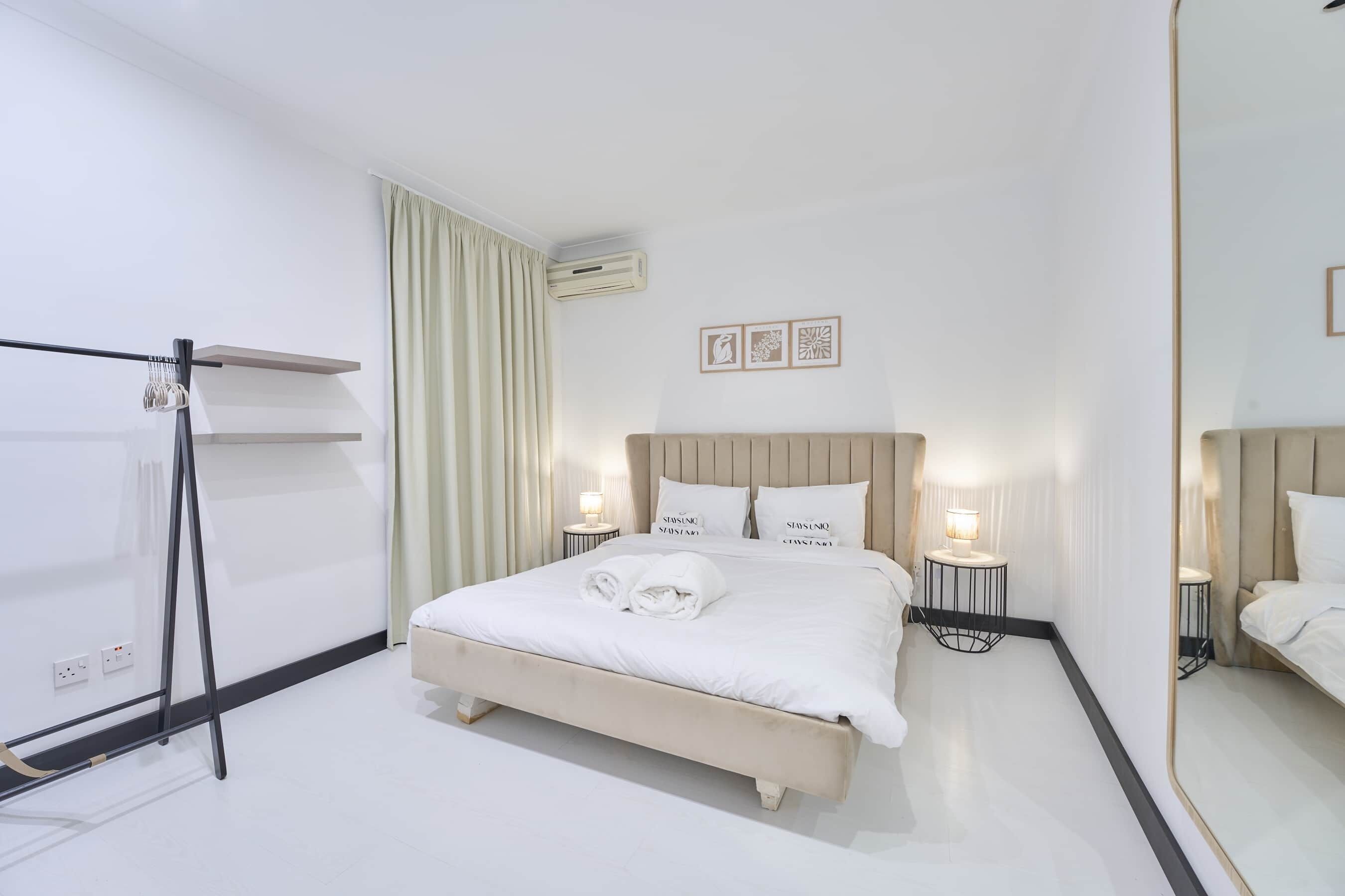 A modern hotel room with a large bed, two bedside tables with lamps, a mirror, and a window with curtains.