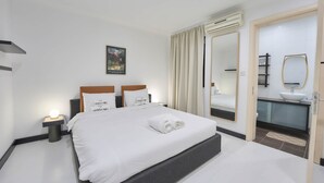 Une chambre d’hôtel moderne dotée d’un grand lit, de tables de chevet avec des lampes, d’une salle de bain avec un lavabo et un miroir, et d’une porte donnant sur une autre pièce.