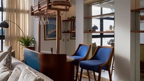 Un salon moderne avec un canapé, deux fauteuils bleus, une table basse en bois, un lampadaire et une grande fenêtre donnant sur un beau paysage.