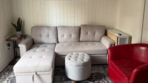 A living room with a sectional sofa, a red chair, a grey ottoman, a small table with a plant, and a painting of a tree on the wall.