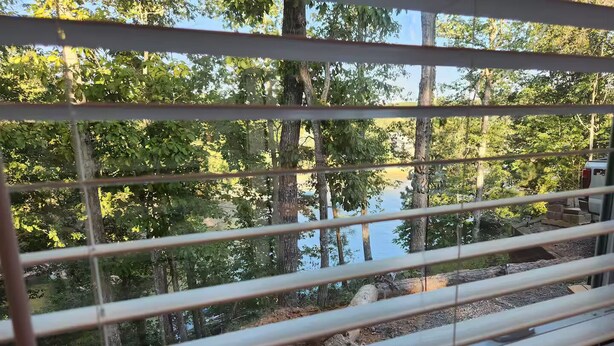 A view through window blinds of a forested area with a body of water and a vehicle in the background.