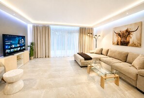 A modern living room with a large flat-screen TV, a beige sofa, a glass coffee table, and a large window with curtains.