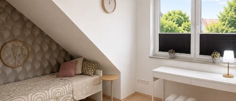 A bedroom with a sloped ceiling, a bed with patterned bedding, a bedside table, a round clock, a window with a view of greenery, and a small stool.