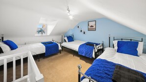 A dormitory room with three beds, each with blue bedding, white sheets, and a black throw blanket. The room has a sloped ceiling, a window with a view, and a small plant on the windowsill. - Contractor 3-Bed Long-Stay & Discounts  (Reading)