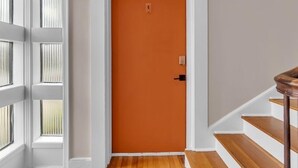 Un couloir avec une porte blanche, un escalier en bois et une fenêtre avec des stores.