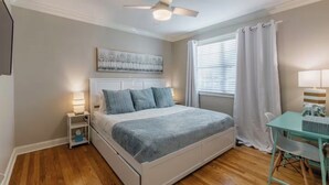 Une chambre à coucher comprenant un lit, des tables de chevet, un ventilateur de plafond, une fenêtre avec des rideaux et une petite table avec des chaises.