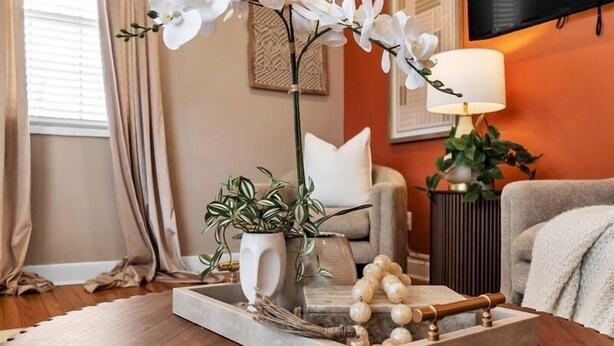 Un salon avec une table basse en bois, une plante d’orchidée blanche, une plante en pot aux feuilles panachées, un plateau décoratif avec des raisins et un téléviseur fixé au mur.
