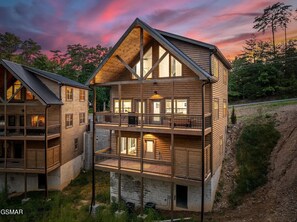 Una baita a più piani in legno con un balcone al secondo piano, immersa tra alberi e un sentiero sterrato.