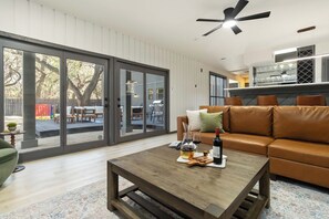 A modern living room with a brown leather sofa, a wooden coffee table with wine glasses and snacks, and a sliding glass door leading to an outdoor patio. - PrivateParadise Heated Pool +HotTub +Sauna +Arcade (San Antonio)
