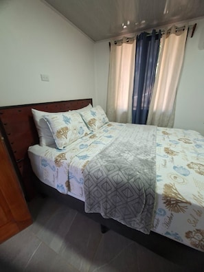 A single bed with a patterned bedspread, a wooden headboard, and a window with curtains.