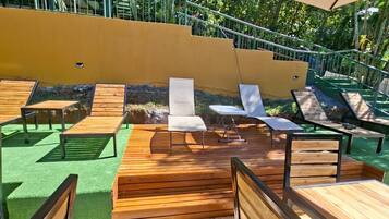 Un área de estar al aire libre con bancos y mesas de madera, rodeada por un muro amarillo y follaje verde.