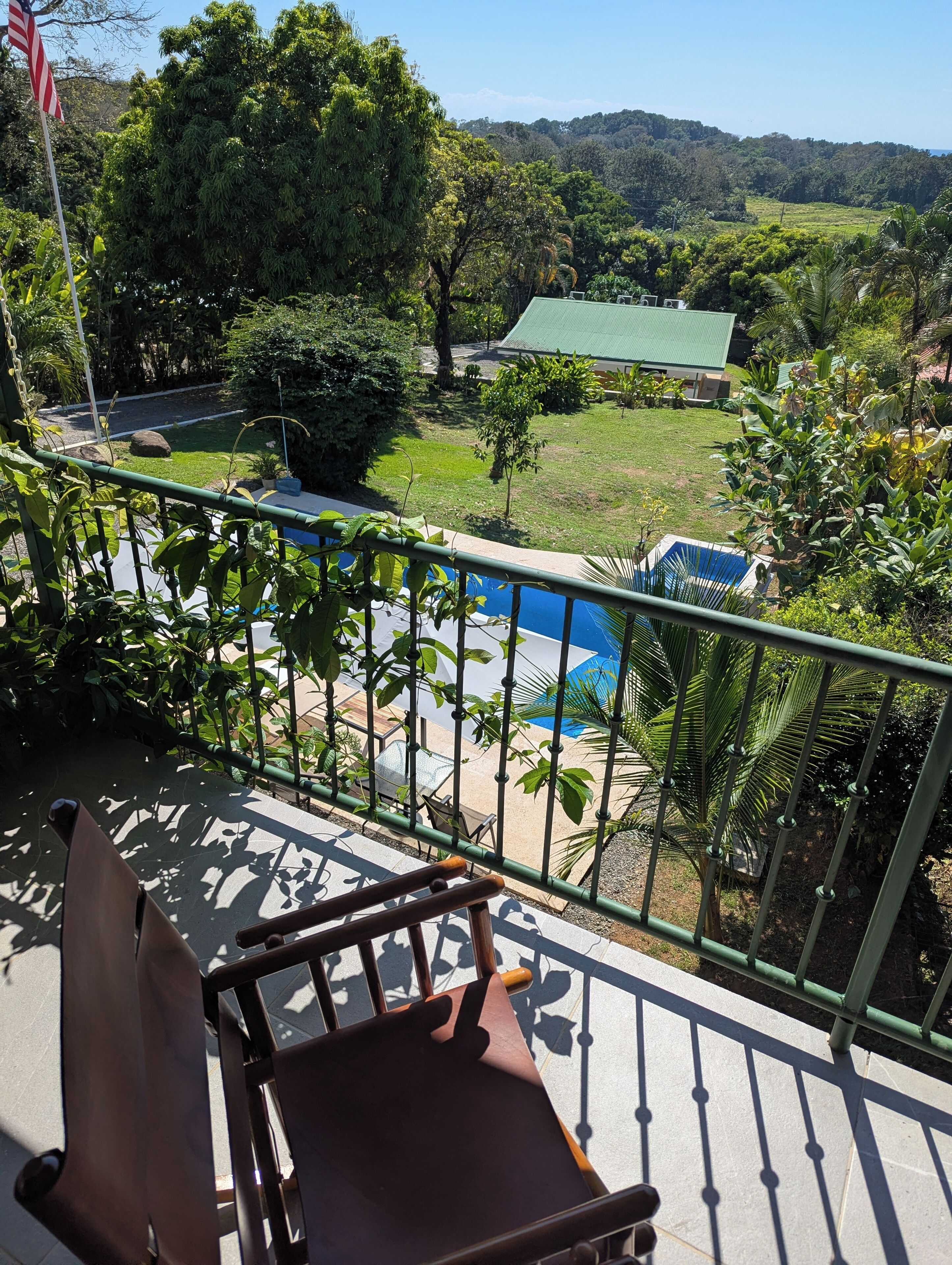 A balcony with a view of a swimming pool, lush greenery, and a distant building.