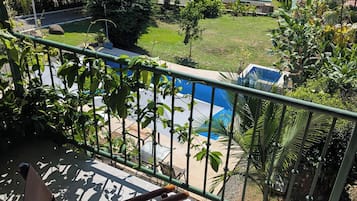 A balcony with a view of a swimming pool, lush greenery, and a distant building.