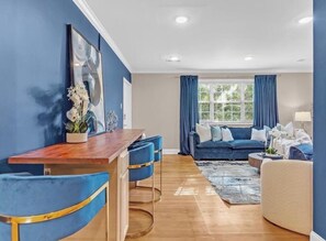 Un salon moderne avec un canapé bleu, une table à manger en bois et des tabourets de bar, ainsi qu’une grande fenêtre avec des rideaux.