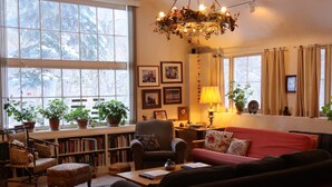 Ein gemütliches Wohnzimmer mit Kamin, Bücherregalen, einem roten Sofa und einem großen Fenster mit Blick auf Bäume.
