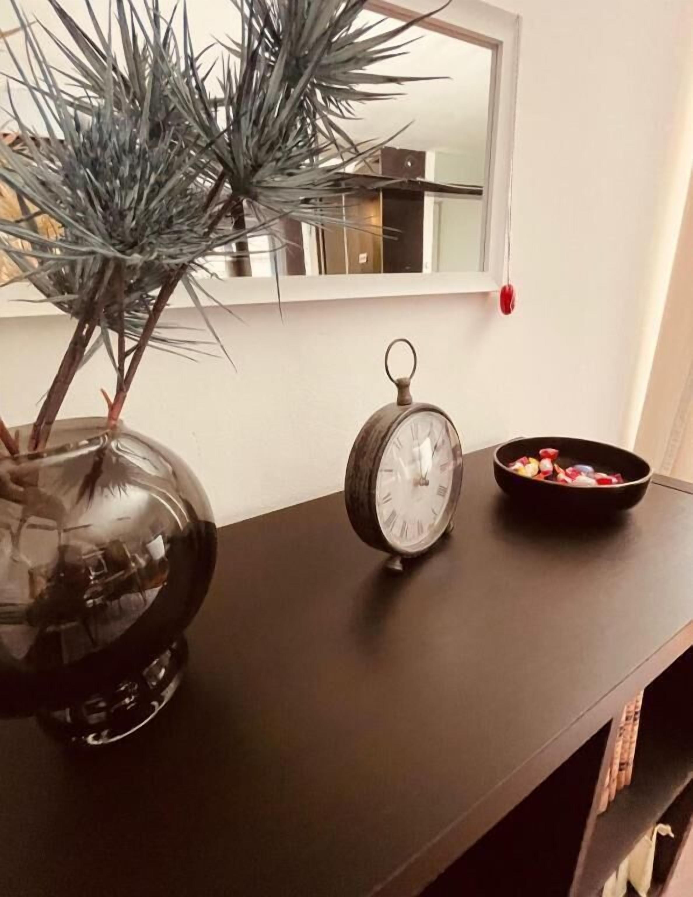 A dark wooden console table with a clear glass vase containing a plant, a vintage-style alarm clock, and a bowl of fruit.