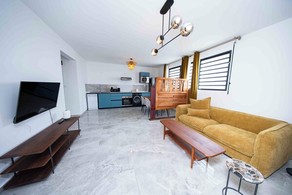 A living room with a sofa, TV, and a wooden bench. A kitchen is visible in the background with a microwave and a washing machine. - Guyane Appart'Hôtel (Cayenne)