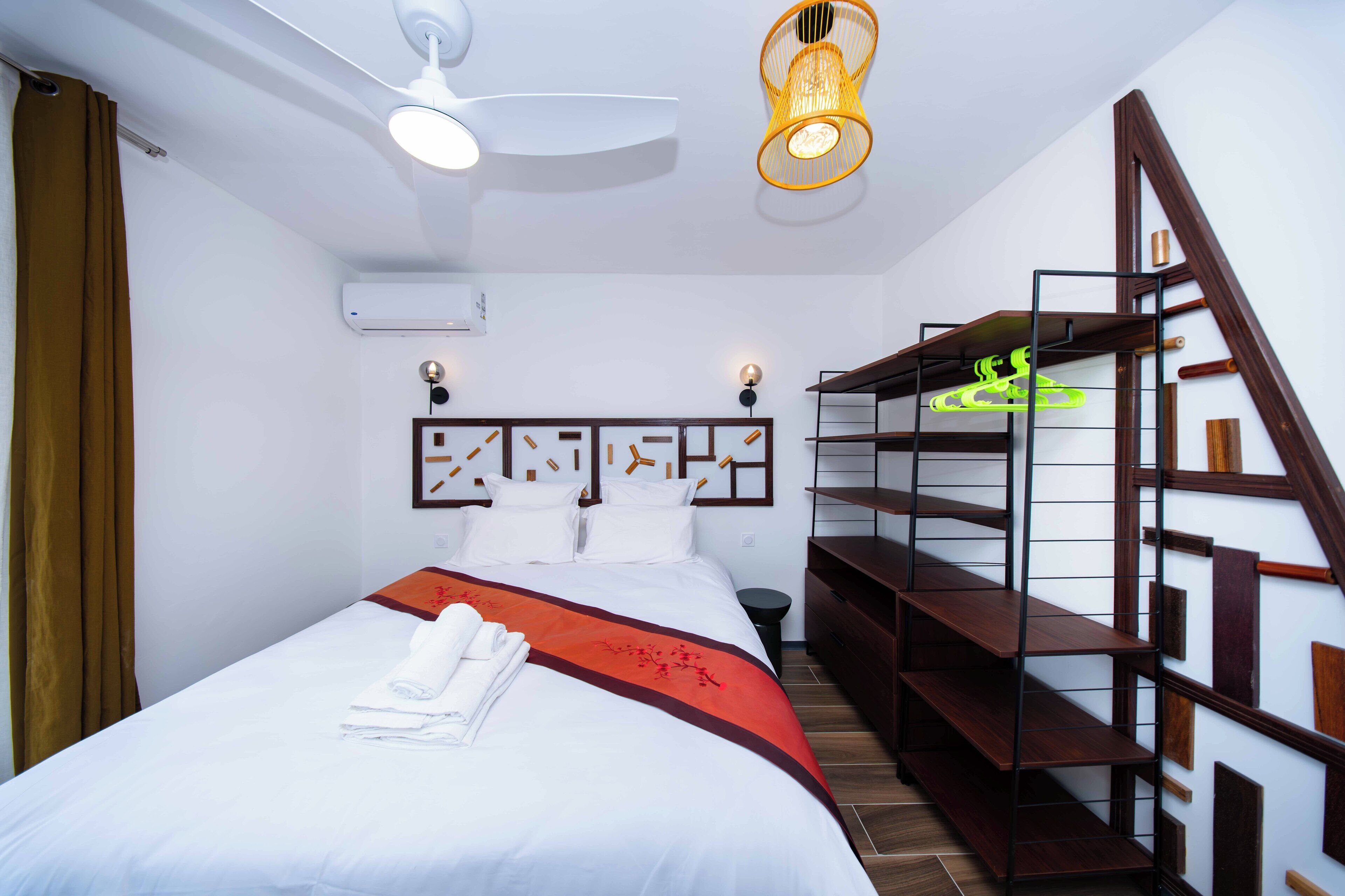Una habitación de hotel con una cama, un ventilador de techo, un estante en la pared y una escalera.