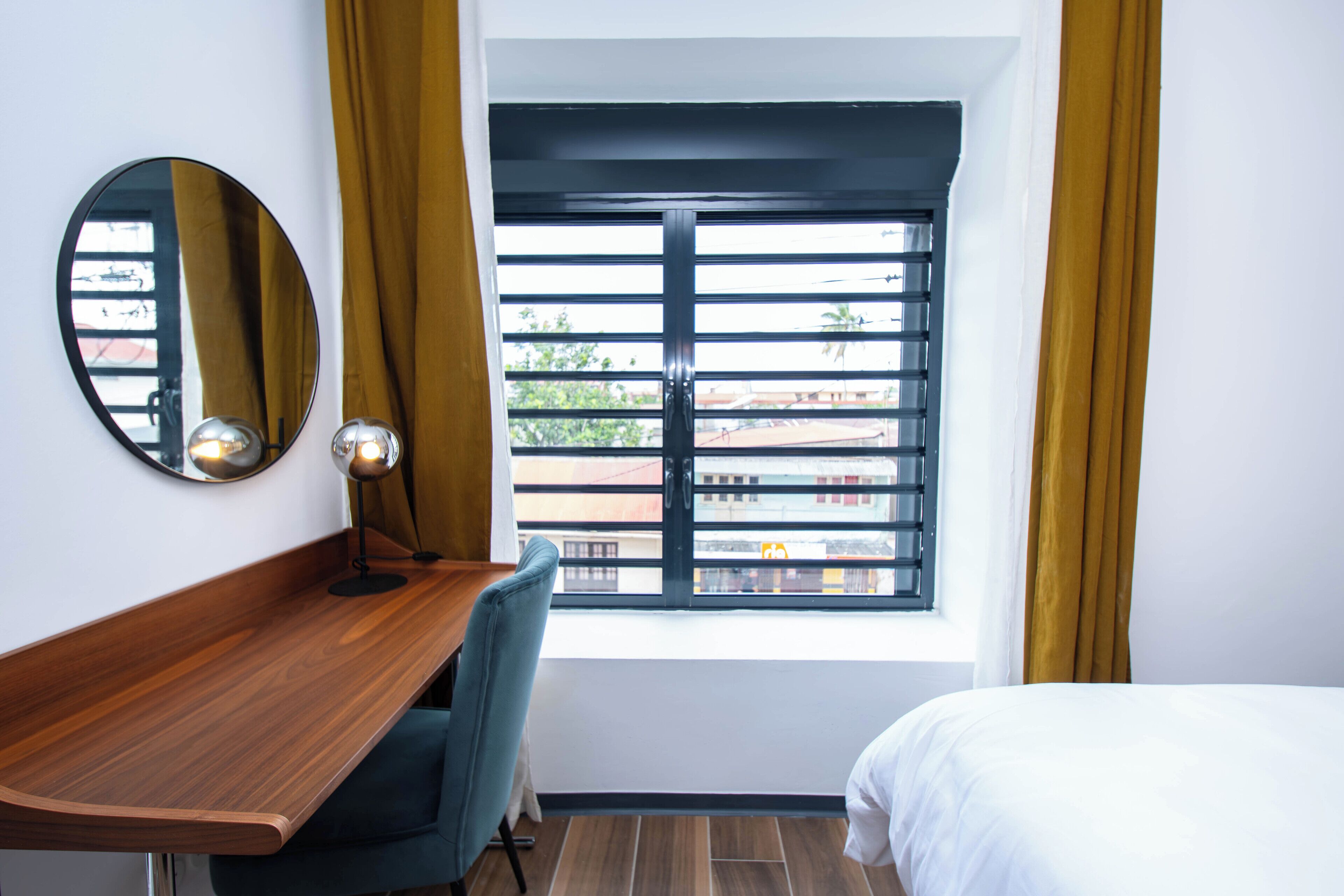 Une chambre d’hôtel avec un bureau en bois, une chaise, un lit, une fenêtre avec des stores noirs et un miroir au mur.