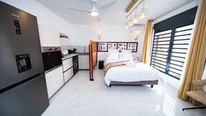 A modern hotel room with a bed, a kitchenette, a ceiling fan, and a large window with curtains.