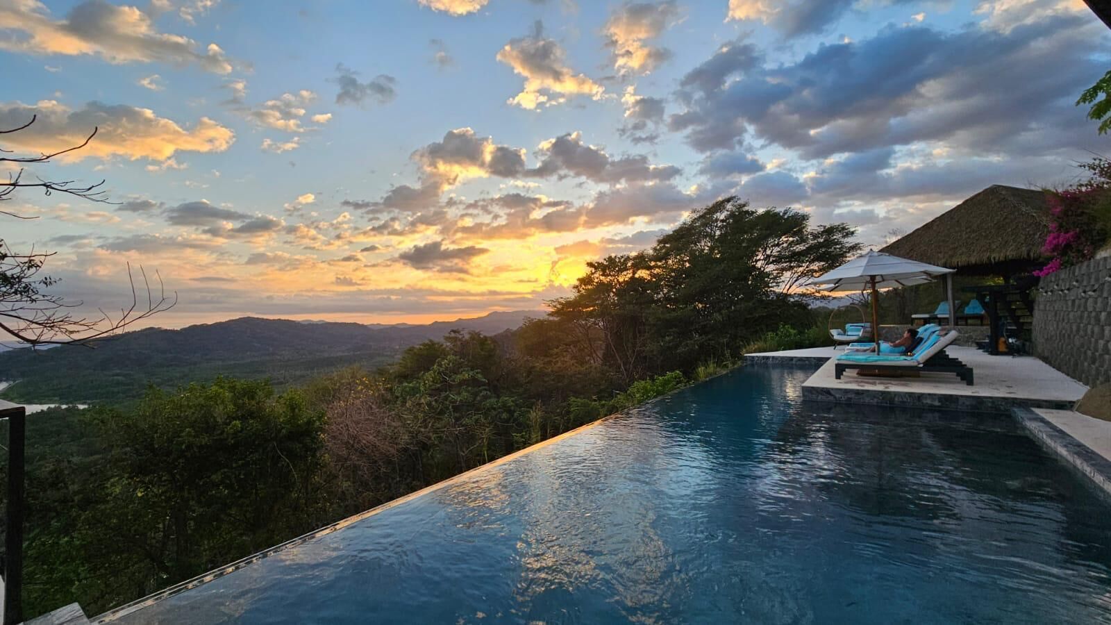 A swimming pool with a view of mountains and a sunset. There are lounge chairs and a thatched-roof structure.