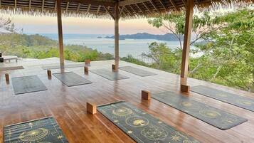 An outdoor wooden deck with yoga mats and meditation cushions, overlooking a scenic coastal view.