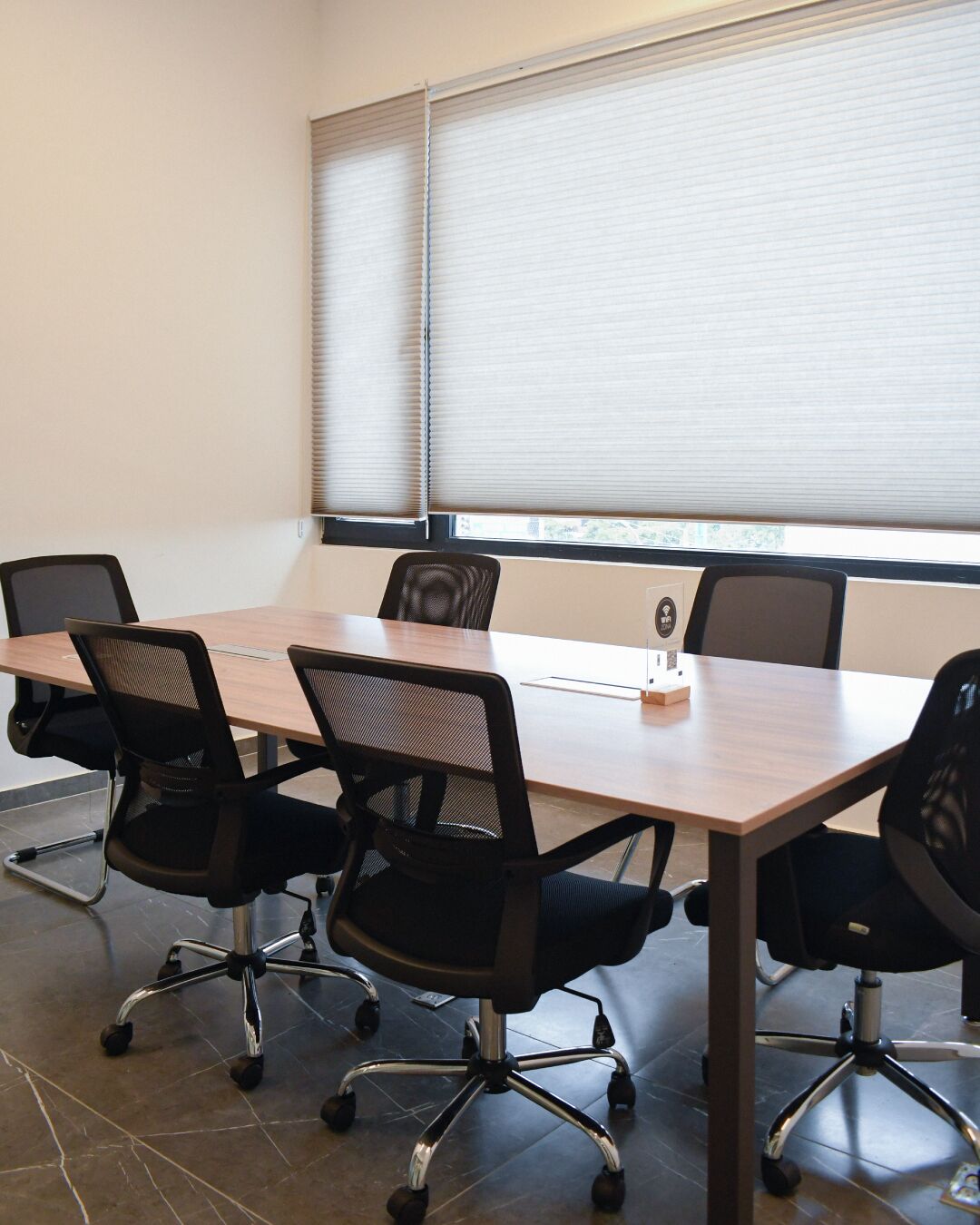 A conference room with a long table, six black office chairs, and a window with blinds.
