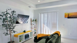 Un salon moderne avec un téléviseur à écran plat, un canapé, une plante en pot, une étagère en bois en forme d’échelle et une grande fenêtre avec des stores.