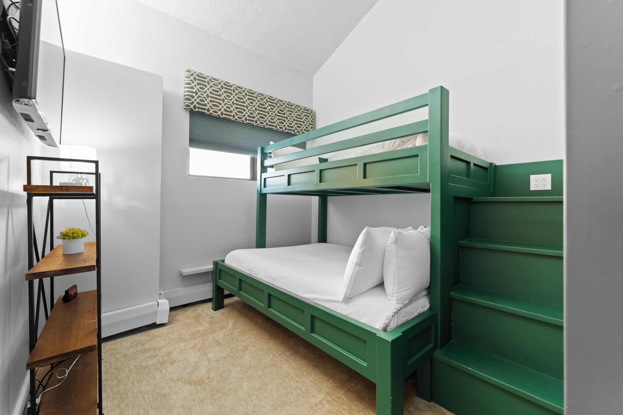 A bunk bed room with a desk and shelves, a window with blinds, and a radiator.