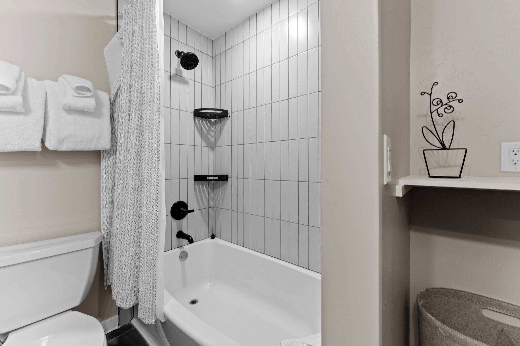 A bathroom with a white bathtub, a shower, and a toilet. There are towels hanging on the wall and a potted plant on a shelf.