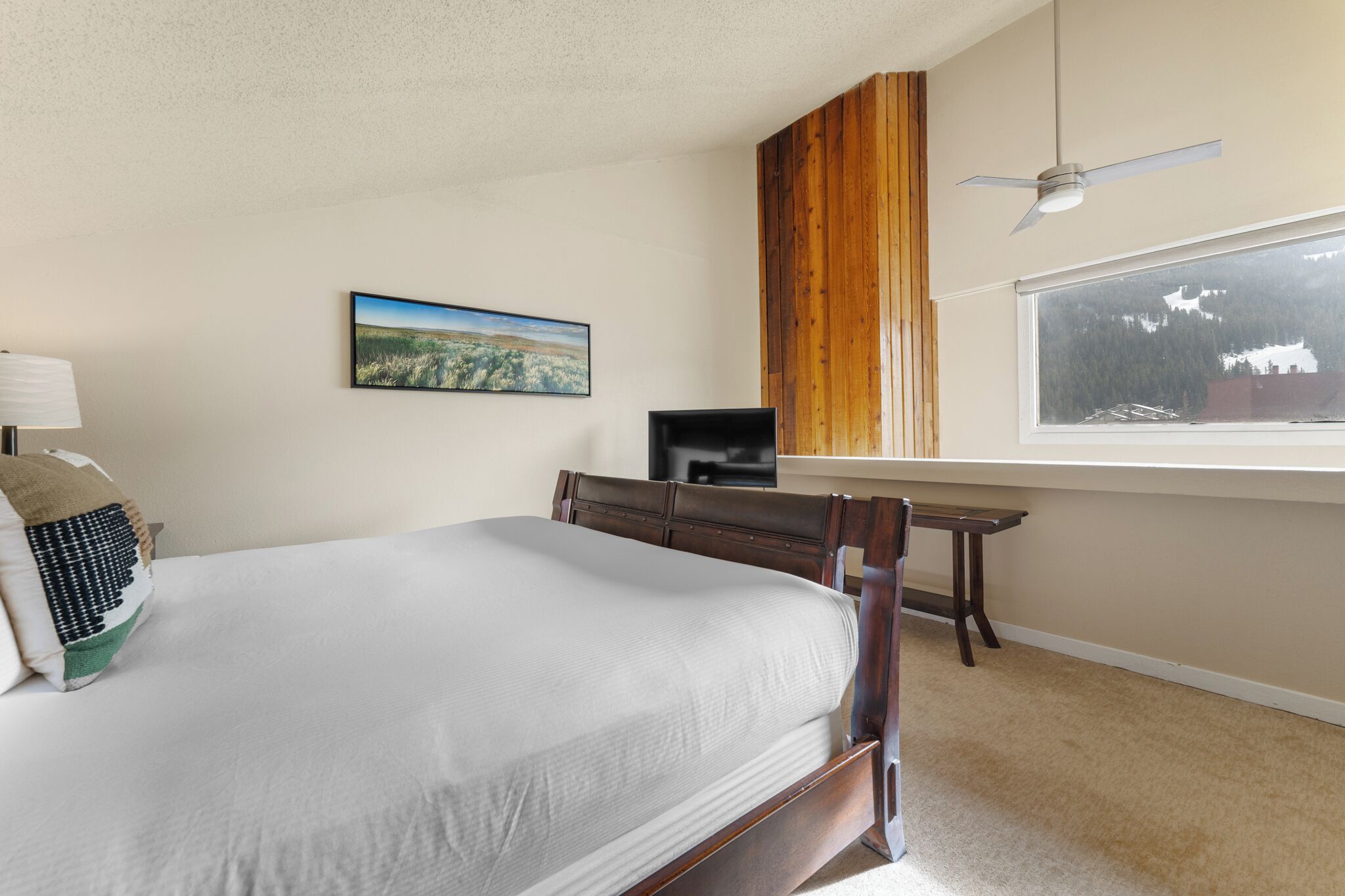 A bedroom with a bed, a desk, a chair, a ceiling fan, a window with a view, and a painting on the wall.