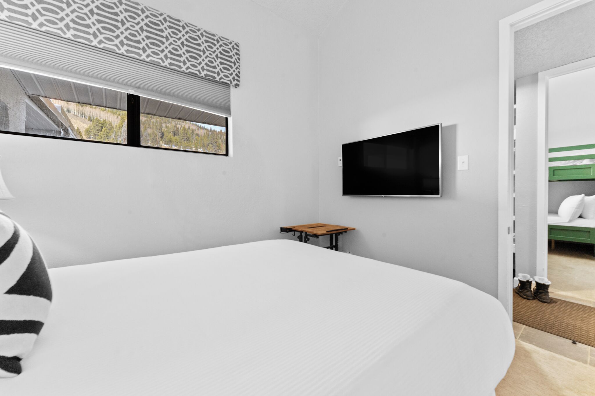 A modern hotel room with a large bed, a small table, a TV mounted on the wall, and a window with a view of trees and mountains.