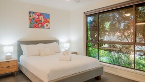 A bedroom with a large bed, two bedside tables, a nightstand, a window with a view of greenery, and a colorful painting on the wall. - Relaxing Family Escape 3-Bedroom Home in Oxley Vale (Oxley Vale)