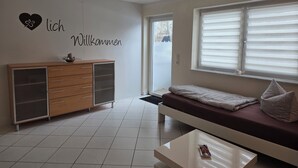 A modern living room with a sofa, a coffee table, a cabinet, and a window with blinds. - Vacation apartment garden view (Schwarzach am Main)