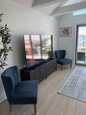 A modern living room with a large flat-screen TV on a black entertainment unit, two matching blue armchairs, a wooden floor, and a view of a residential area through a large window. - Spacious 4-bedroom house in enchanting Atlantic City with AC (Atlantic City)