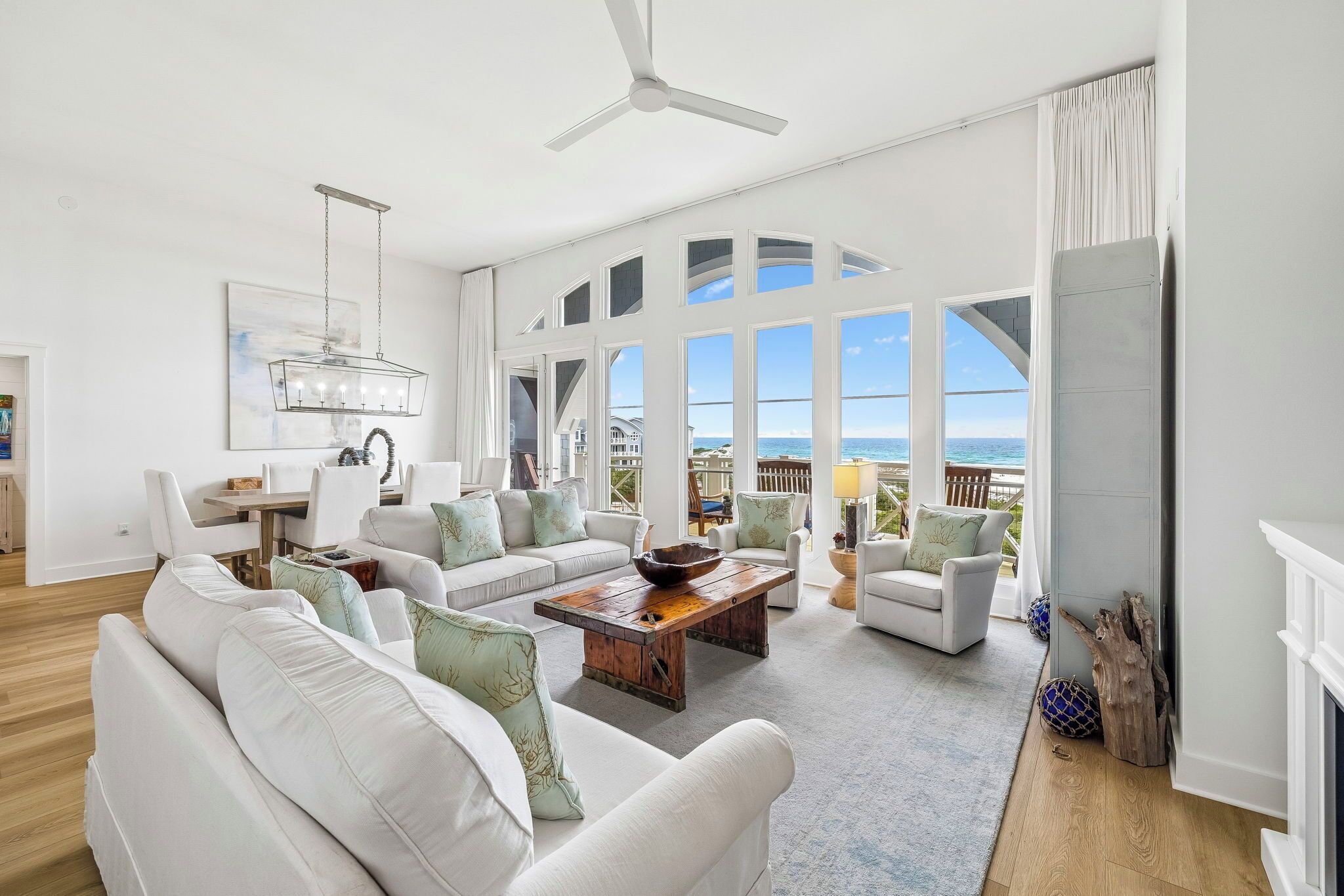 A spacious living room with white furniture, a wooden coffee table, and large windows offering ocean views.