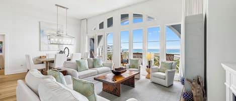 A spacious living room with white furniture, a wooden coffee table, and large windows offering ocean views.