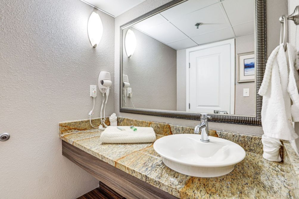 Une salle de bain avec un comptoir en granit, un lavabo blanc et un grand miroir. On y trouve des sèche-cheveux fixés au mur et une serviette accrochée à la porte.